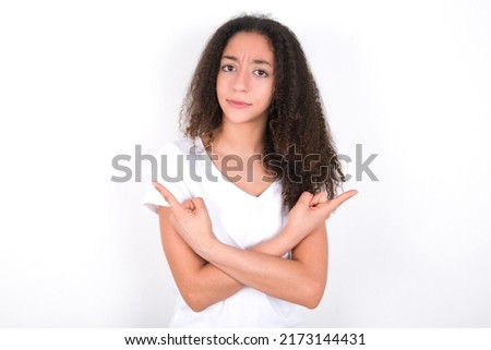 Serious Teenager girl with afro hair style wearing white t-shirt over white background crosses hands and points at different sides hesitates between two items. Hard decision concept