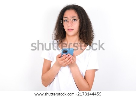 Photo of astonished crazy Teenager girl with afro hair style wearing white t-shirt over white background hold smartphone dislike feedback concept