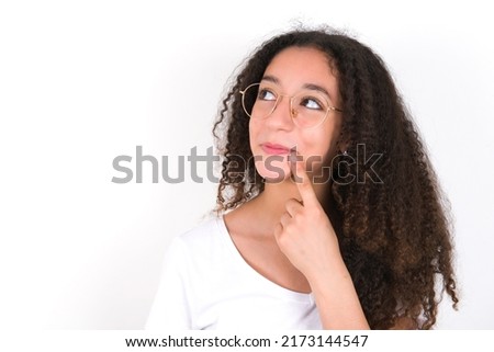 Lovely dreamy Teenager girl with afro hair style wearing white t-shirt over white background keeps finger near lips looks aside copy space. Royalty-Free Stock Photo #2173144547