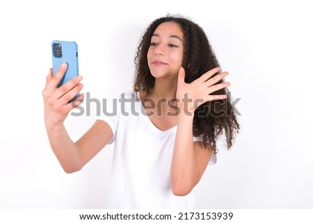 Teenager girl with afro hair style wearing white t-shirt over white background holds modern mobile phone and makes video call waves palm in hello gesture. People modern technology concept