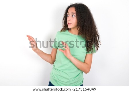 Displeased Teenager girl with afro hair style wearing green t-shirt over white background keeps hands towards empty space and asks not come closer sees something unpleasant