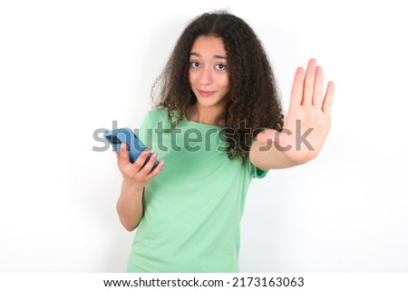 Teenager girl with afro hair style wearing green t-shirt over white background using and texting with smartphone with open hand doing stop sign with serious and confident expression, defense gesture
