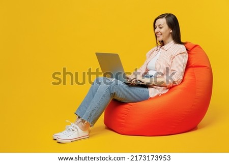 Full body young happy woman she 30s wears striped shirt white t-shirt sit in bag chair hold use work on laptop pc computer isolated on plain yellow background studio portrait. People lifestyle concept