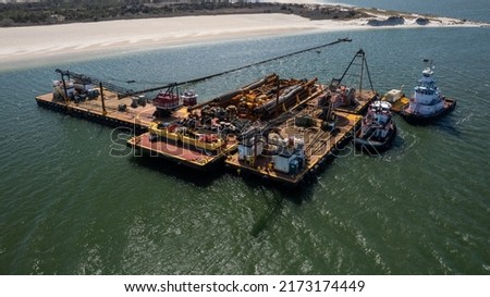 An aerial shot of three barges with construction equipment on them, pushed together by two tugboats. It is a sunny day and the water is calm on Long Island, NY.