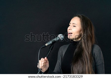 Young Asian or Caucasian brunette woman in casual clothes holding mic and singing song alone in music studio on black background. Recording voice to self-made composed album. Hobby, education