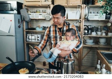 japanese asian new dad holding a smartphone is taking a photo with his cute baby girl in arm while cooking breakfast in the kitchen at home