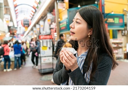 closeup shot of asian woman visitor having baked doughnut and looking into space trying to find her way out at the exit of kuromon ichiba market in Osaka japan