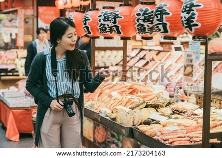 asian female tourist pointing at fresh seafood displayed in a shop selling aquatic products in kuromon ichiba market in Osaka japan. translation on lantern: âseafoodâ and âsushiâ Royalty-Free Stock Photo #2173204163