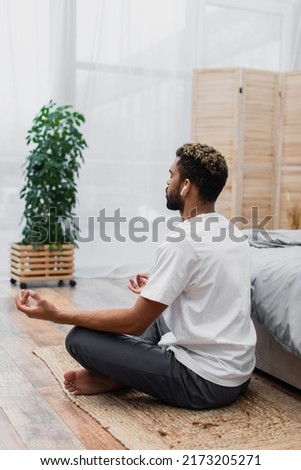 bearded african american man in wireless earphone meditating while sitting near bed at home