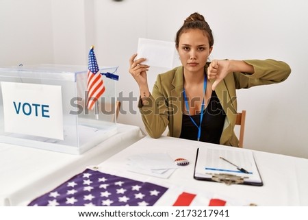 Beautiful hispanic woman holding voting envelop in ballot box with angry face, negative sign showing dislike with thumbs down, rejection concept 