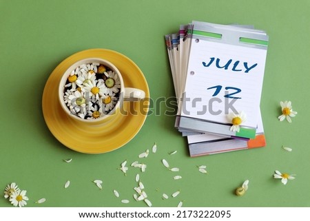 Calendar for July 12: numbers 12 and the name of the month July on a sheet calendar, next to a cup of chamomile tea, pastel background, top view.