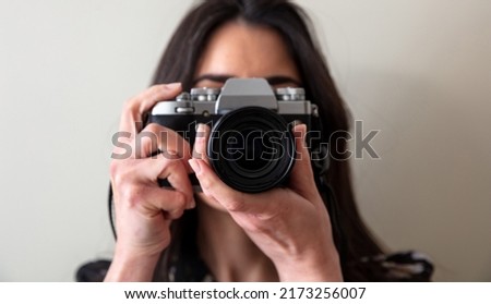 Young woman holding photo camera is shooting your picture. Girl photographer focus at you, close up view