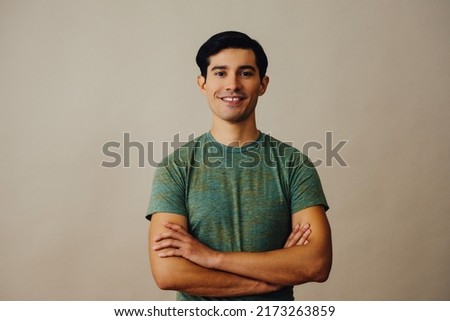 Portrait hispanic latino man with arms crossed and black hair smiling handsome young adult green t-shirt over gray background looking at camera studio shot Royalty-Free Stock Photo #2173263859