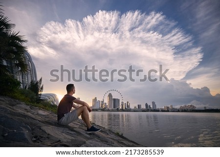 Young man sitting on waterfront and looking at dramatic clouds of thunderstorm above urban skyline, Singapore.
 Royalty-Free Stock Photo #2173285539
