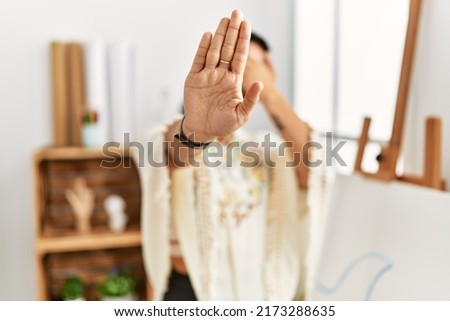 Young hispanic woman with short hair at art studio covering eyes with hands and doing stop gesture with sad and fear expression. embarrassed and negative concept. 