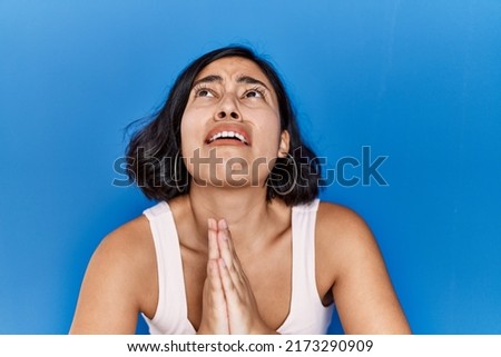Young hispanic woman standing over blue background begging and praying with hands together with hope expression on face very emotional and worried. begging. 