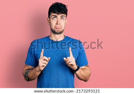 Young hispanic man wearing casual t shirt pointing up looking sad and upset, indicating direction with fingers, unhappy and depressed. 