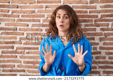 Beautiful brunette woman standing over bricks wall moving away hands palms showing refusal and denial with afraid and disgusting expression. stop and forbidden. 