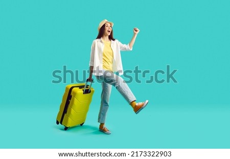 Happy woman with yellow suitcase going on summer holiday. Smiling overjoyed young girl wearing jeans, shirt and panama hat walking with her travel bag on bright turquoise background. Vacation concept Royalty-Free Stock Photo #2173322903