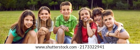 Happy children sitting on green grass and smiling. Cheerful little kids enjoying their school break, spending time in a summer camp, making new friends, and playing outside. Group portrait. Banner Royalty-Free Stock Photo #2173322941