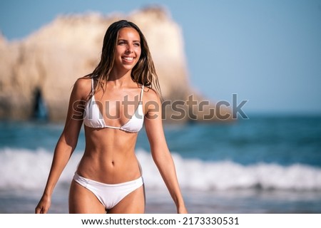 fit young woman in white swimwear walking in wavy sea and looking away while spending summer vacation on beach in Algarve