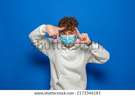 Young caucasian man with curly hair wearing a medical mask standing over isolated blue background making a finger frame with his hands. Concept of creativity and photography.