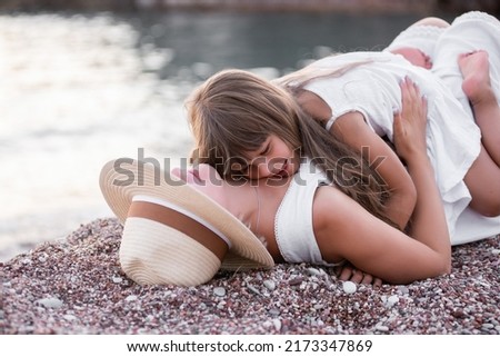 Millennial mother with daughter fooling around, having fun on the sand of the beach at sea. Little girl hugs middle-aged woman in hat, tickles mom. The journey of single parent. Travel trip pregnancy
