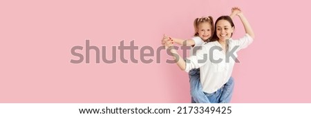Mom woman in light clothes has fun with cute baby girl. Little baby daughter sitting on mother's back isolated on pastel pink background. Web banner.