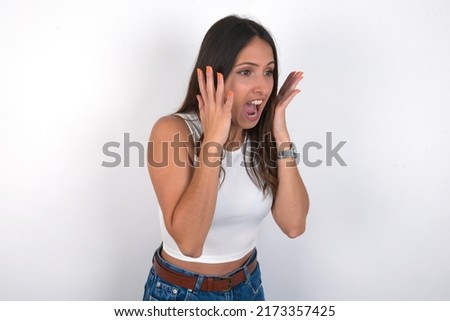 young beautiful caucasian woman wearing white top over white background  with shocked facial expression, holding hands near face, screaming and looking sideways at something amazing.