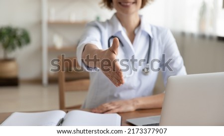 Close up smiling woman doctor extending hand for handshake to camera, physician therapist gp wearing white uniform with stethoscope sitting at work desk, greeting patient at medical appointment Royalty-Free Stock Photo #2173377907