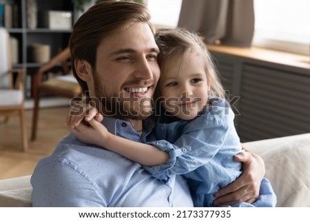 Head shot cute small baby girl cuddling neck of smiling young father, enjoying calm sweet tender moment together relaxing on sofa, watching TV series or cartoons in living room, holiday pastime.