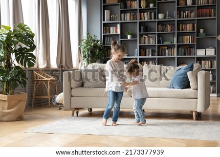 Full length loving small girl holding hands of small toddler sister, dancing barefoot together in floor carpet in living room. Happy two little cute children involved in energetic activity indoors. Royalty-Free Stock Photo #2173377939