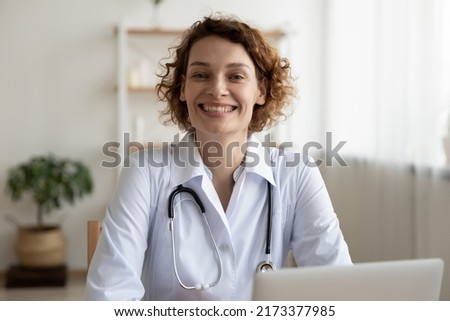 Head shot portrait smiling woman doctor sitting at work desk with laptop in hospital, friendly young nurse physician gp wearing white uniform with stethoscope looking at camera, healthcare concept
