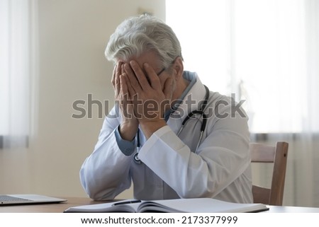 Stressed frustrated mature doctor wearing white uniform with stethoscope covering face with hands, sitting at work desk in hospital, sad senior gp feeling desperate and guilty for medical failure Royalty-Free Stock Photo #2173377999