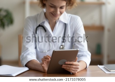 Close up smiling woman doctor using digital tablet, medical apps, sitting at desk in hospital, young nurse therapist physician wearing white uniform working online, consulting patient