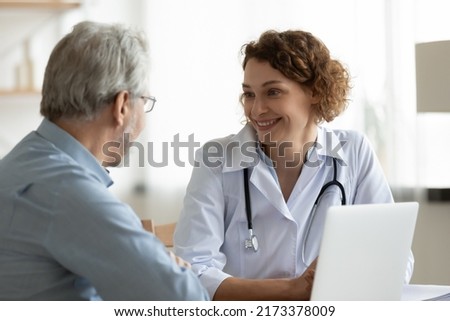 Smiling woman doctor listening to mature patient complaints at meeting in hospital, friendly therapist physician wearing white uniform with stethoscope consulting senior man at medical appointment Royalty-Free Stock Photo #2173378009