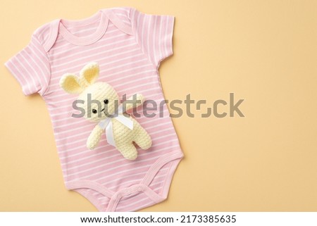 Baby accessories concept. Top view photo of knitted bunny toy over pink bodysuit on isolated pastel beige background