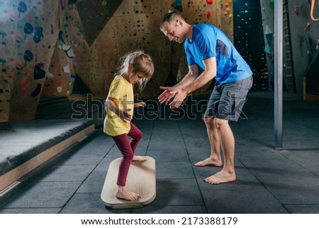 Father supporting his daughter while she training on fitness balance board in a gym. Little girl using exercise accessory rocker board. Family sport activities. Rock climbing wall on background.