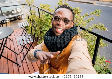 young african man outdoors, sitting outside cafe happy smiling takes selfie making v sign with hand, looking at camera, perspective from phone.