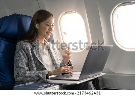 Attractive asian female passenger of airplane sitting in comfortable seat listening music in earphones while working at modern laptop computer with mock up area using wireless connection on board. Royalty-Free Stock Photo #2173462431