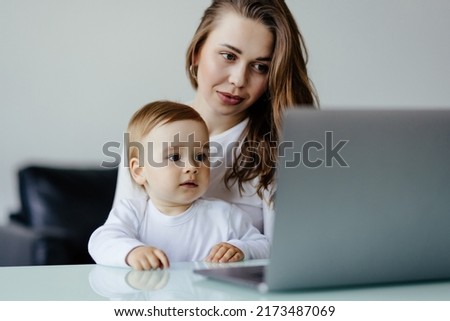 Young single mother mom with toddler newborn baby infant son using laptop, searching web, watching cartoons together.