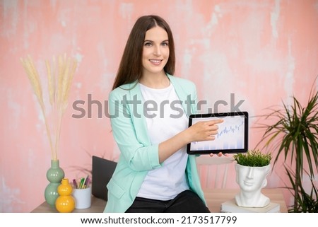 Happy girl holding tablet with financial graphic and showing at it in colorful office
