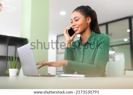 Charming young African-American woman using laptop on the workplace and has phone conversation, female office worker chatting on the smartphone while typing on a laptop