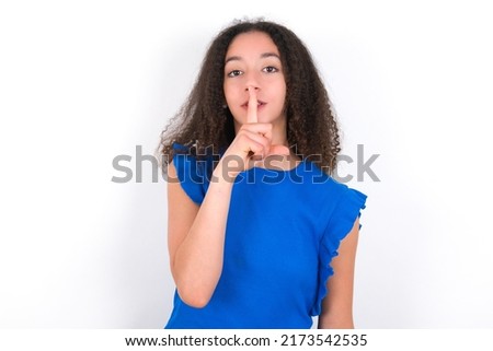 Teenager girl with afro hair style wearing blue t-shirt over white background makes silence gesture, keeps finger over lips. Silence and secret concept.