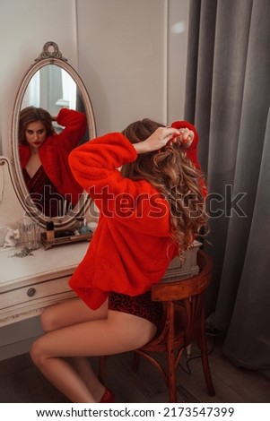 teen party. young glamour girl in red fur coat is sitting near mirror in bedroom and tying red ribbon in hairstyle near curtain on white wall background. valentine's day, lifestyle concept, free space