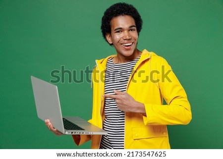 Jubilant fun young black curly man 20s wears yellow waterproof raincoat outerwear hold use work on laptop pc computer pointing index finger on screen isolated on plain green background studio portrait