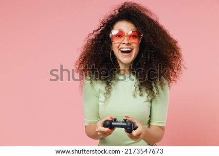 Gambling happy overjoyed fancy young curly latin woman 20s wears mint t-shirt sunglasses hold in hand play pc game with joystick console isolated on plain pastel light pink background studio portrait