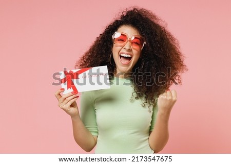 Excited young curly latin woman 20s wears mint t-shirt sunglasses hold gift certificate coupon voucher card for store do winner gesture isolated on plain pastel light pink background studio portrait