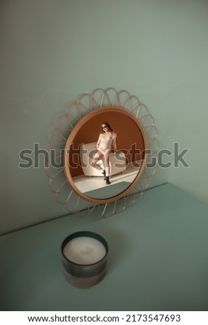 round mirror on mint wall background. young pregnant girl in beige bodysuit is sitting casual with hand on belly on white chair near brown wall in reflection of mirror. pregnant concept, free space