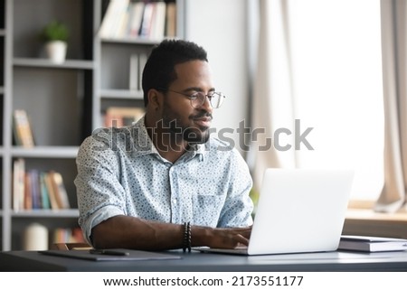 Satisfied African American man wearing glasses looking at computer screen, reading good news in email, chatting in social network with friends, freelancer blogger working on online project Royalty-Free Stock Photo #2173551177